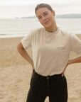 Model wearing Temple Cycles X Clara Jonas women’s graphic tee in cream, beach setting.