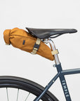 Waterproof roll-top cycling bag in burnt orange attached to a bike saddle, studio setting.