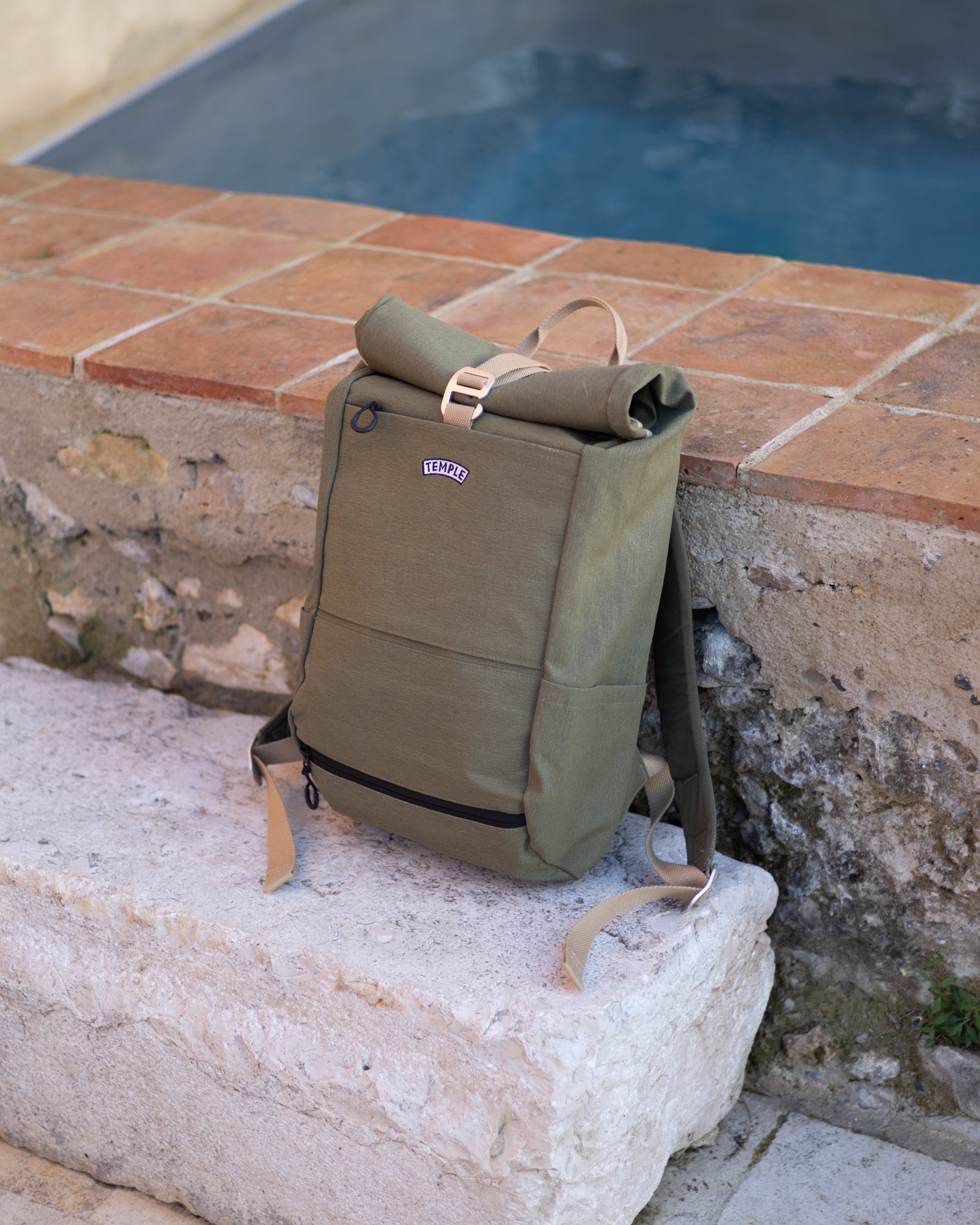 Temple Cycles water-resistant roll-top pannier backpack in olive green, resting on stone ledge beside a pool.