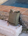Temple Cycles water-resistant roll-top pannier backpack in olive green, resting on stone ledge beside a pool.