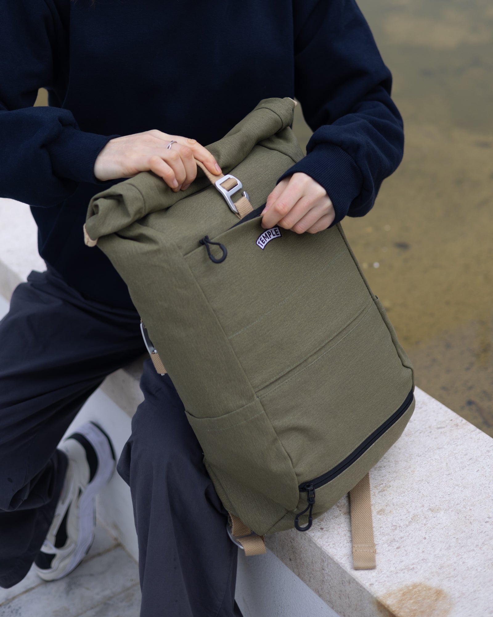 Close-up of a person opening Temple Cycles olive green weather-proof roll-top pannier backpack while sitting down outside.