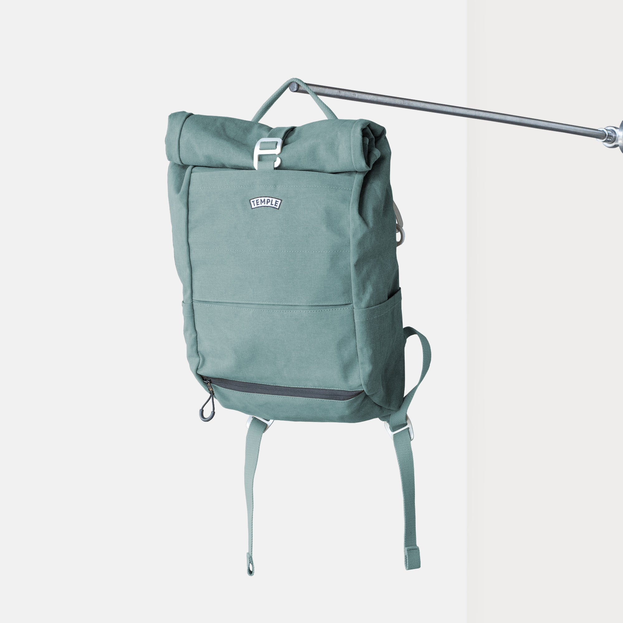 Temple Cycles roll-top backpack in Stonewash Teal canvas with side pockets, and secure buckle closure hanging in a studio setting.