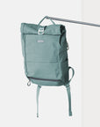 Temple Cycles roll-top backpack in Stonewash Teal canvas with side pockets, and secure buckle closure hanging in a studio setting.