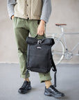 Model holding casual men’s black backpack, urban setting. 