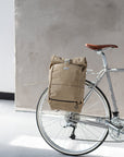 Beige roll-top backpack on rear pannier bike rack, urban setting. 