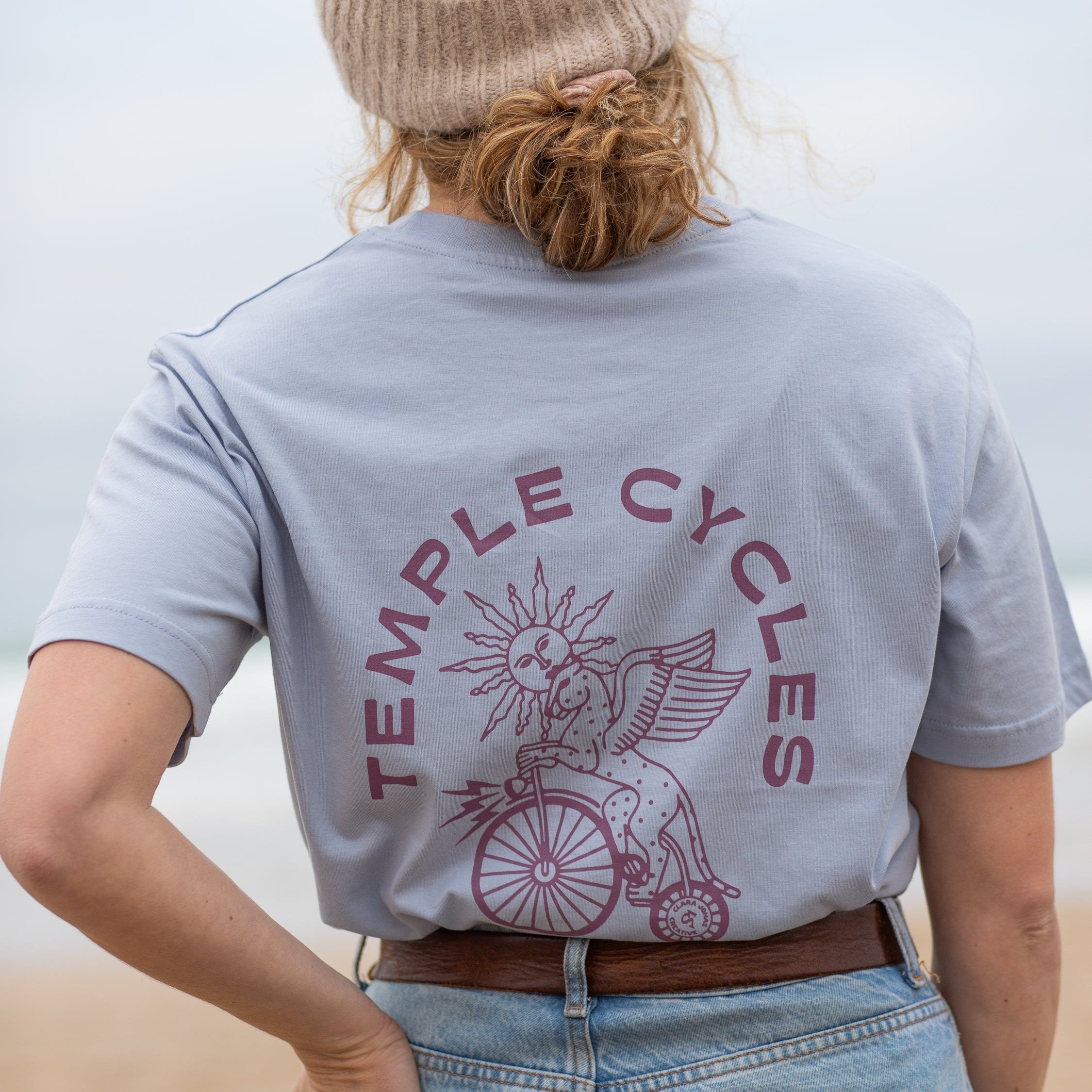 Blue unisex organic cotton T Shirt with purple graphic on female model, beach setting.