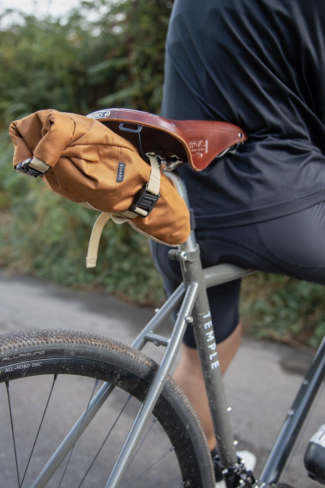 Orange roll-top bike bag attached to leather saddle, rural setting.