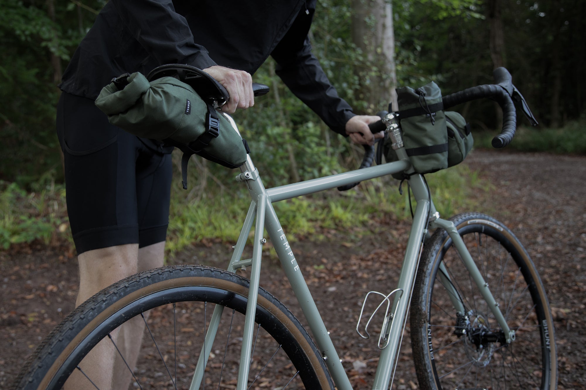 Model holding a touring bike with three green cycling bags attached, rural setting. 