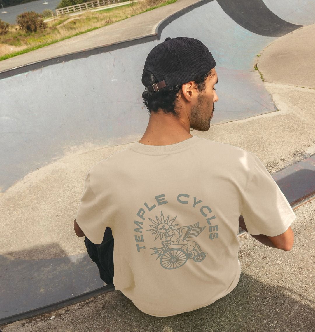 Back view of model wearing supper soft cream and green men’s graphic tee, skate park setting.